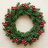 Artificial red berry picks with brown stems are spaced around a green pine wreath on a light beige background.