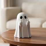 A white ghost figurine with black eyes and raised middle fingers sits on a round wooden table in a softly blurred living room setting.
