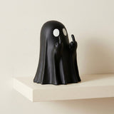 A black ghost figurine with white circular eyes and raised middle fingers, displayed on a white shelf against a light beige wall.