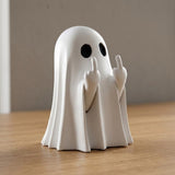 A white ghost figurine with black circular eyes and raised middle fingers, standing on a wooden surface against a neutral gray background.