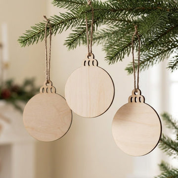 Three round unfinished wood ornament blanks hang from green Christmas tree branches on brown twine against a softly blurred cream background.