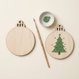 Two round wooden ornament-shaped craft pieces on a cream background, one blank and one painted green Christmas tree, with a paintbrush and a small dish of green paint.