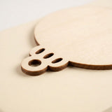 An unfinished wood ornament blank rests on a pale surface, showing its dark edge, hanging hole, and three decorative oval cutouts.