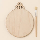 A round unfinished light wood ornament blank with decorative top cutouts lies beside a paintbrush on a cream background.
