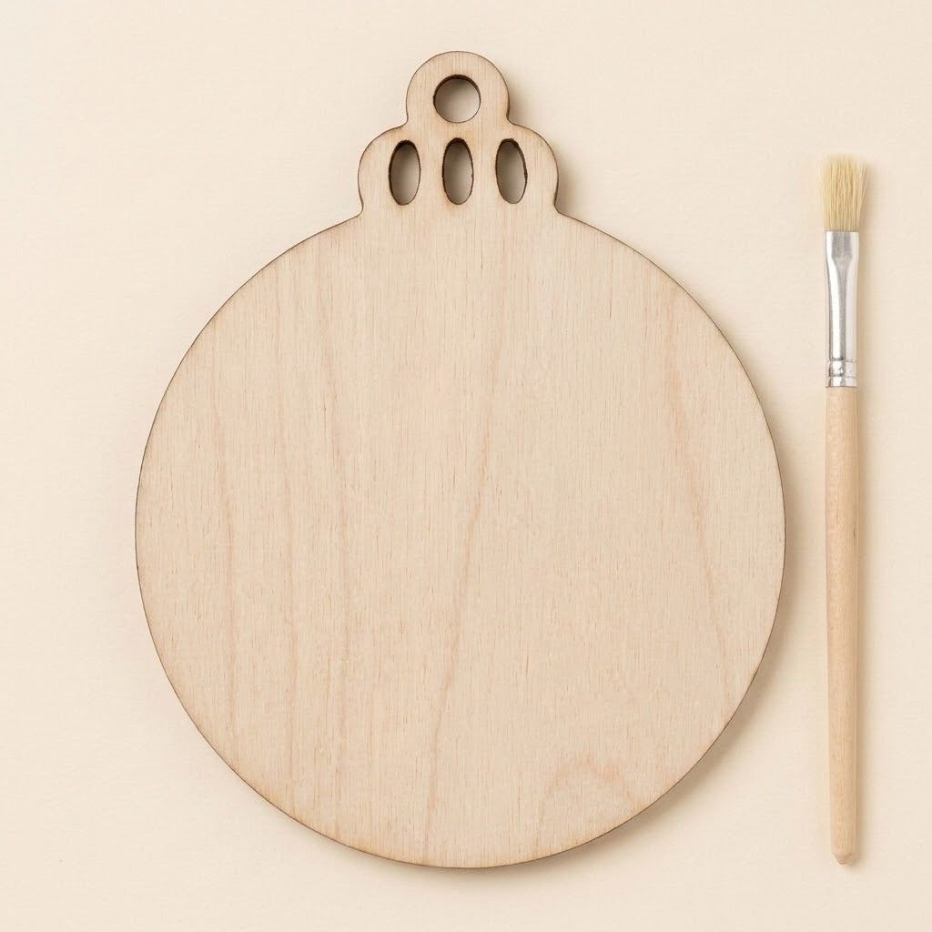 A round unfinished light wood ornament blank with decorative top cutouts lies beside a paintbrush on a cream background.