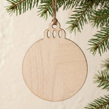 An unfinished light wood ornament blank hangs by brown twine from a green Christmas tree branch against a cream background.