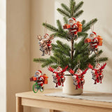 A small evergreen Christmas tree displayed on a light wood table, decorated with multiple hanging Santa Claus ornaments in different poses, including riding bicycles and riding reindeer, with gift boxes and a white pot.