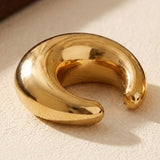 A polished gold-tone ear cuff rests on a textured beige surface, casting a shadow beside its rounded open ends.