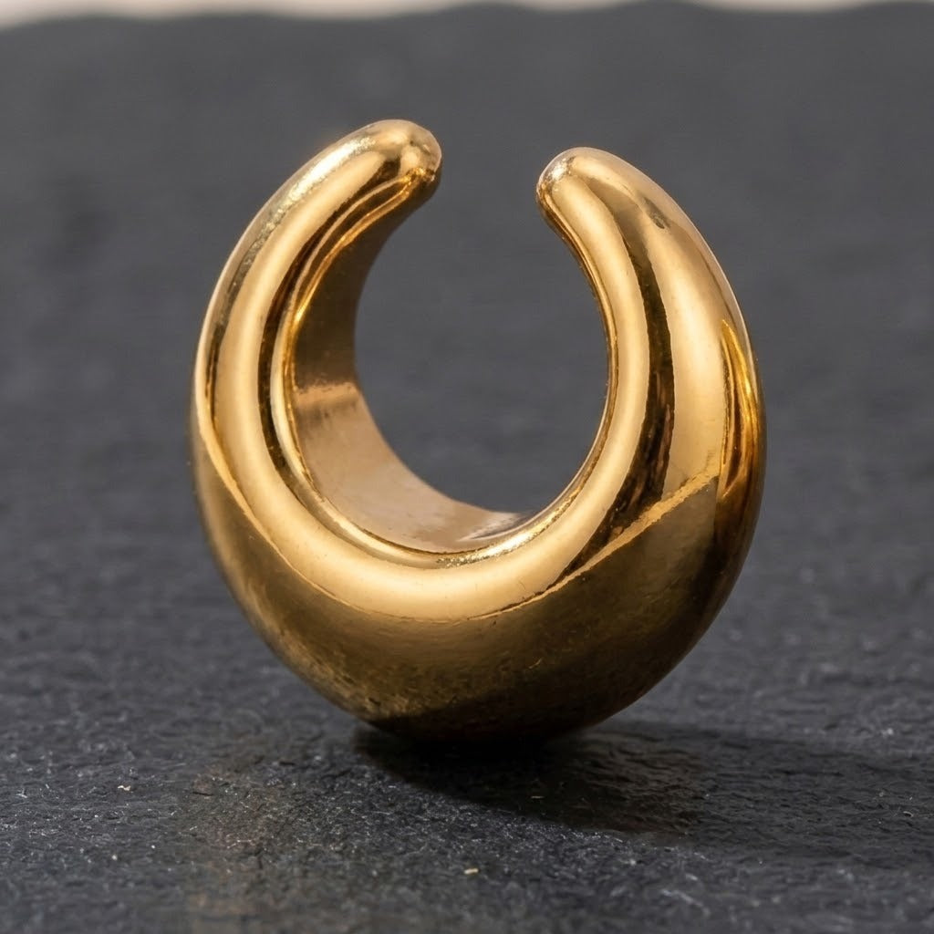 A polished gold crescent-shaped ear cuff or hoop earring, shown standing upright on a dark textured slate surface.