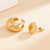 A polished gold-tone ear cuff sits beside a round stud with a visible post and backing on a cream surface.