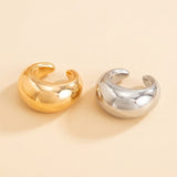 Gold-tone and silver-tone ear cuffs sit side by side on a warm cream background, showing polished surfaces and C-shaped openings.