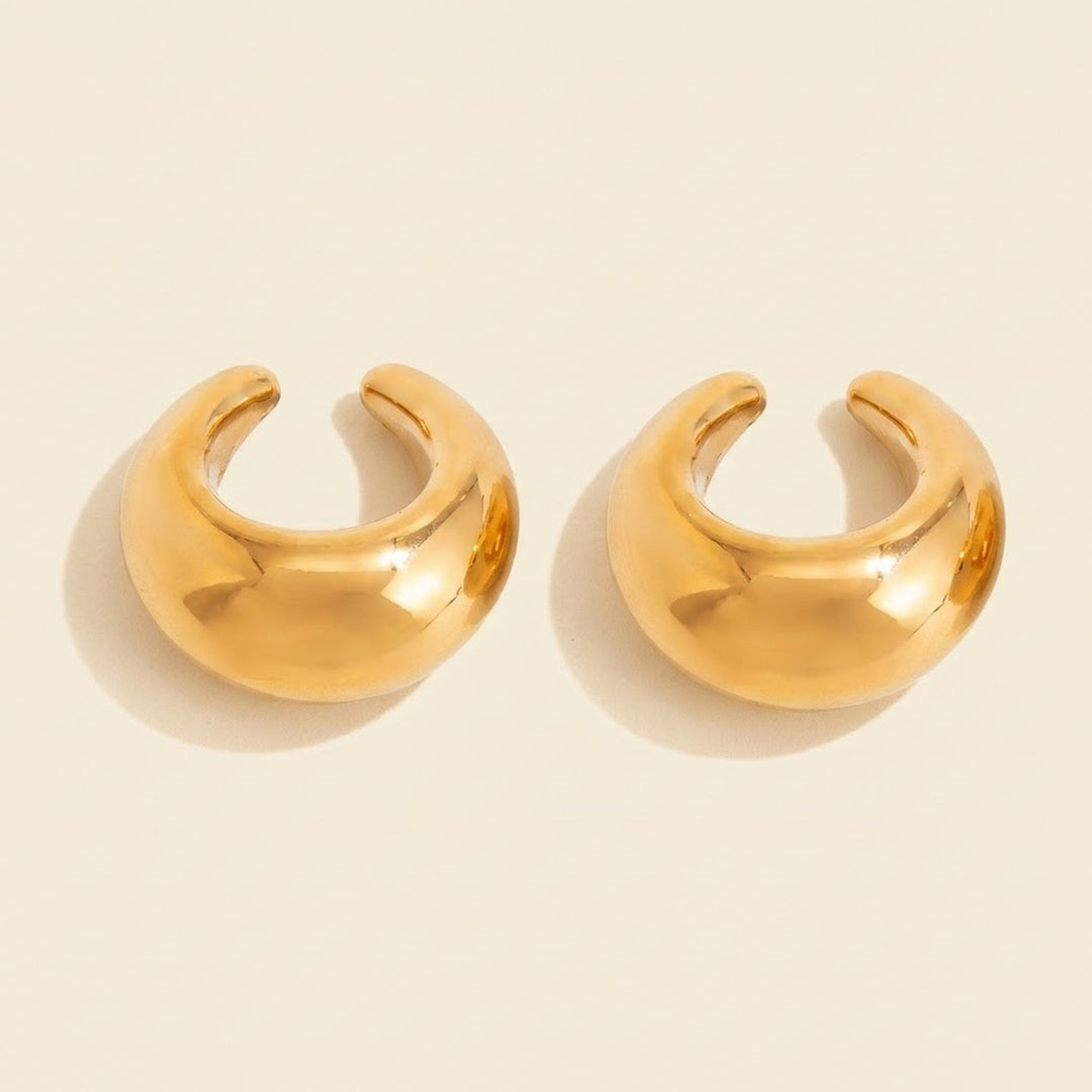 Two polished gold-tone ear cuffs sit on a warm cream background, with C-shaped openings and soft shadows.