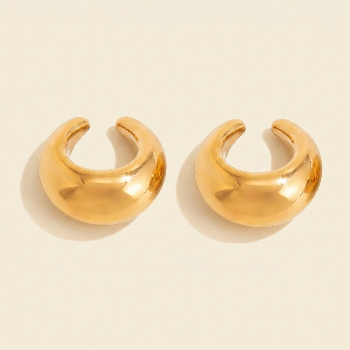 Two polished gold-tone ear cuffs sit on a warm cream background, with C-shaped openings and soft shadows.