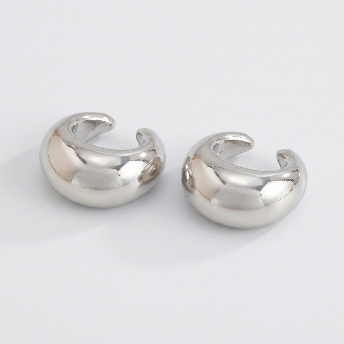 Two polished silver-tone ear cuffs rest on a light gray background, with rounded profiles and open C-shaped designs.