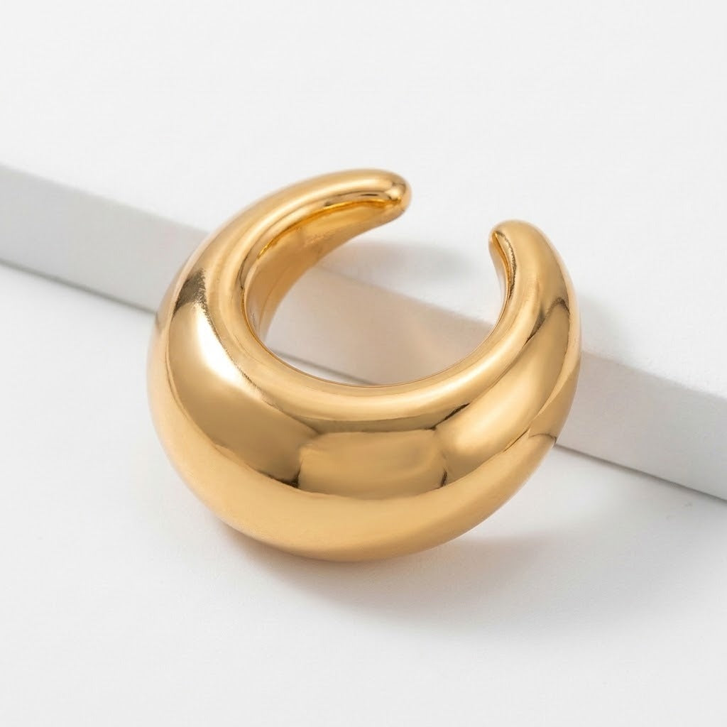 A polished gold-tone ear cuff rests beside a raised white edge, with its C-shaped opening facing upward.