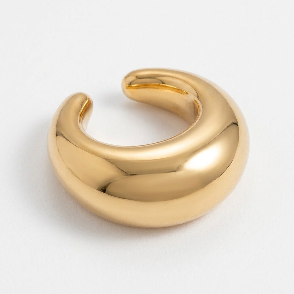 A gold-tone ear cuff rests at an angle on a white background, showing its wide curved face and rounded ends.