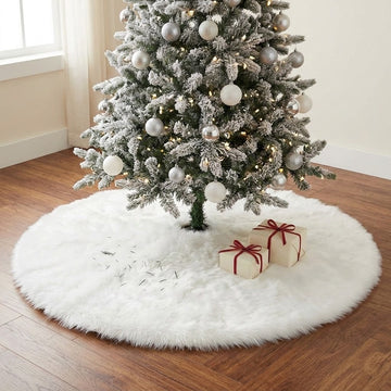 A round white fluffy tree skirt surrounds a frosted Christmas tree, with scattered needles and two wrapped gifts on a wooden floor.