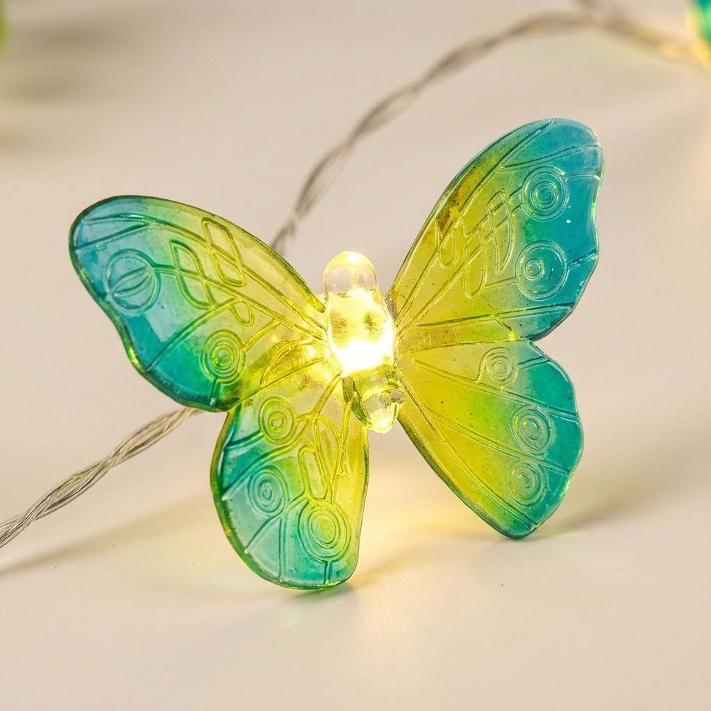 Butterfly string lights shown close up with a glowing warm white LED and translucent yellow and turquoise PVC wings against a cream background.