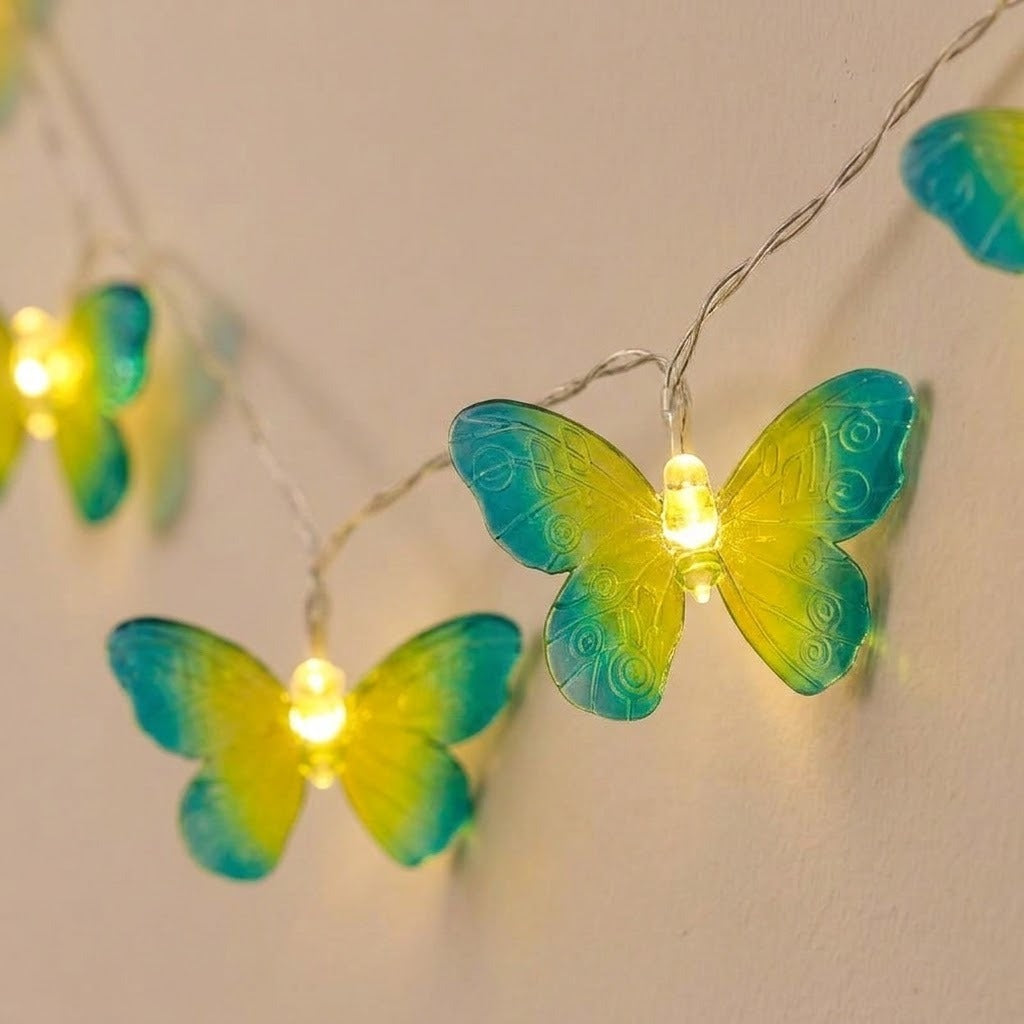Yellow and turquoise butterfly string lights hang from silver wire against a beige wall, with patterned translucent wings.