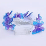 Unlit purple and blue butterfly string lights surround bundled silver wire and a clear battery box on a pale background.