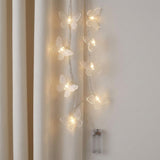 Clear butterfly string lights hang vertically beside a cream curtain, with a transparent battery box against the pale wall.