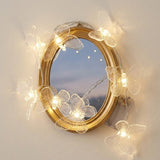 Clear butterfly string lights wrap around an oval gold-framed mirror on a cream wall, with a transparent battery box alongside.