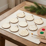 Unfinished wood ornament craft set rests on beige cloth atop a wooden table beside paintbrushes, a paint palette, and evergreen branches.