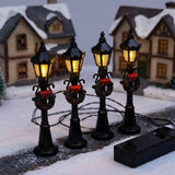 Four black miniature street lights glow yellow beside snowy model houses, with wreaths, red bows, connecting wires, and a battery box.