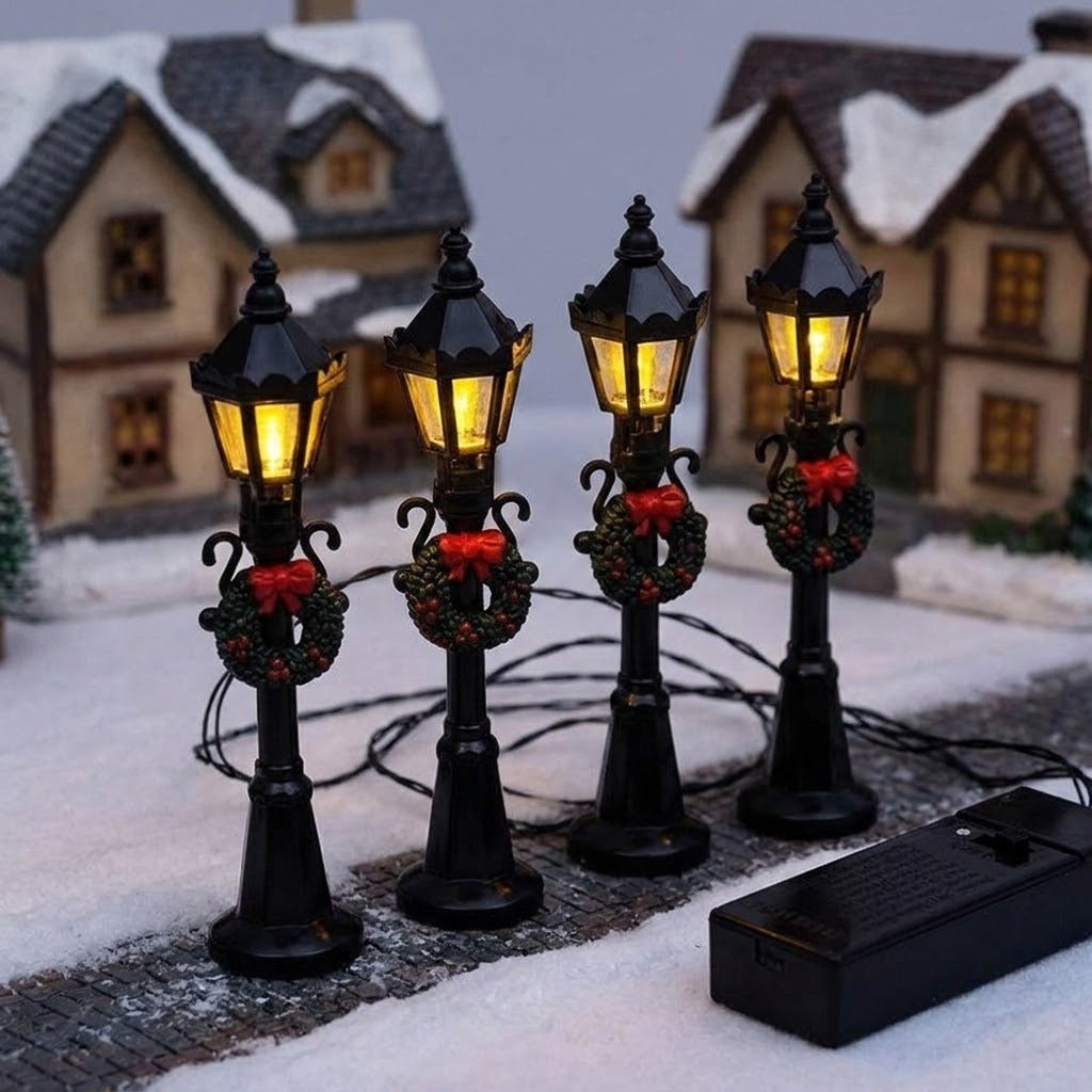 Four black miniature street lights glow yellow beside snowy model houses, with wreaths, red bows, connecting wires, and a battery box.