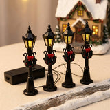 Four black miniature street lights with wreaths and red bows glow yellow beside a snowy model path and Christmas cottage.