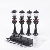Four unlit black miniature street lights with clear lanterns, wreaths, and red bows connect to a battery box against a white background.