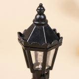 A close-up of one black plastic miniature street light shows its pointed lantern roof, scalloped edging, and clear panels against beige.
