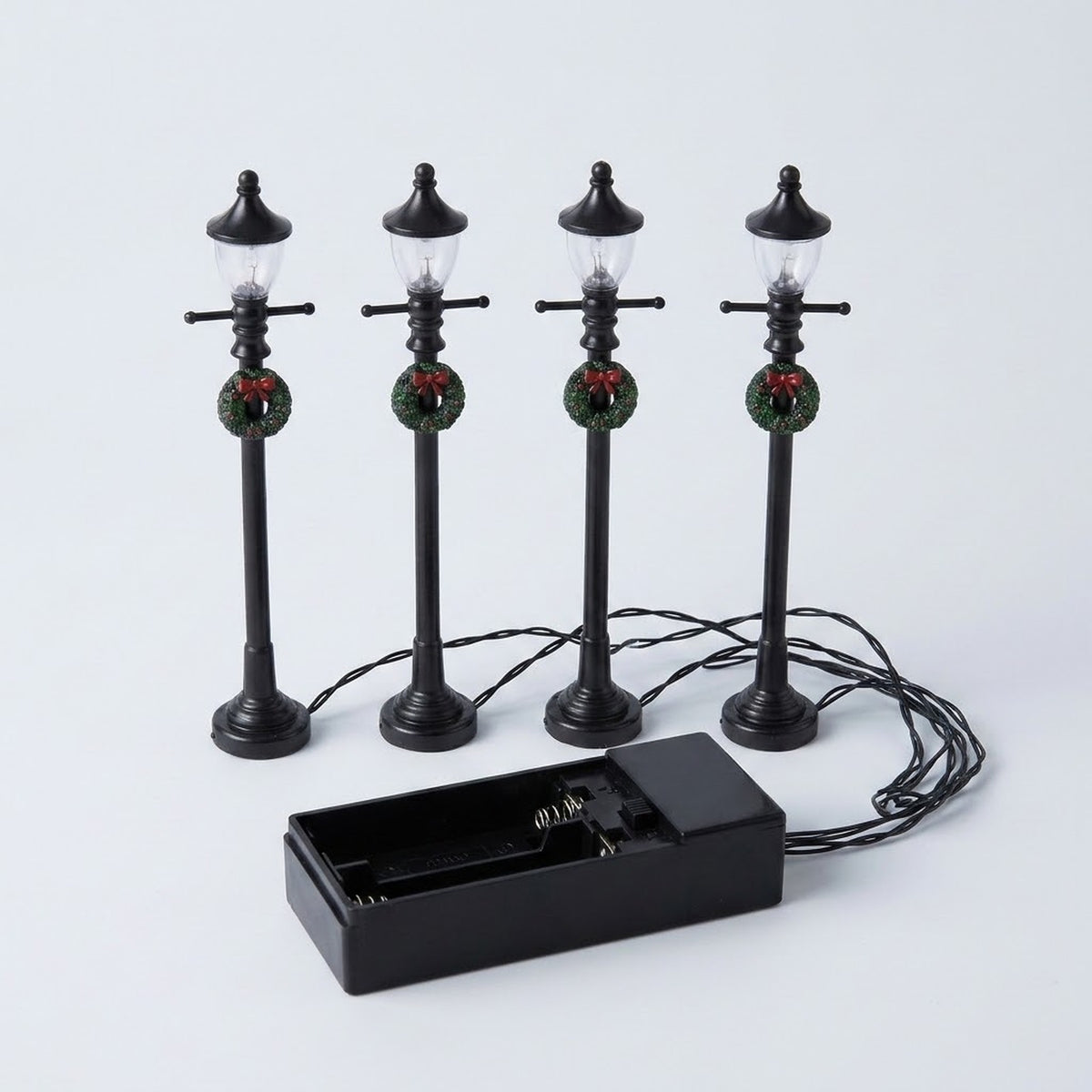 Four black miniature street lights with crossbars, pointed lanterns, and wreaths stand against white behind an open battery box and connecting wires.