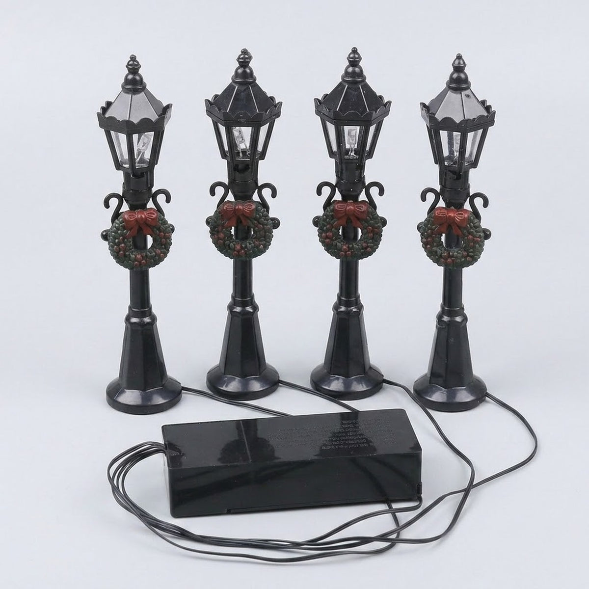 Four black plastic miniature street lights with pointed lanterns, green wreaths, and red bows connect to a battery box on a pale background.