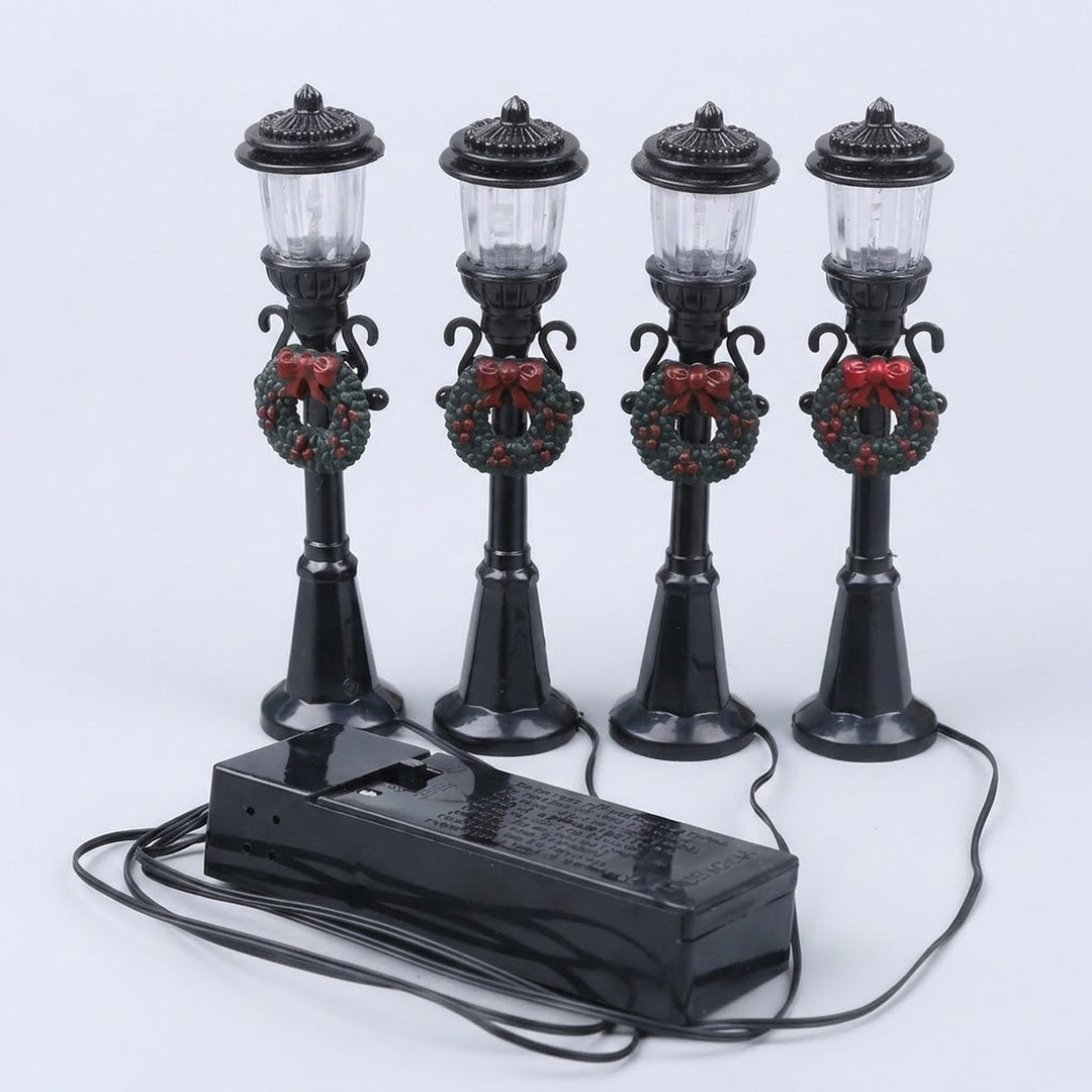 Four black plastic miniature street lights with round lanterns, green wreaths, and red bows stand behind a wired battery box against white.