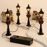 Four glowing black miniature street lights with wreaths and red bows surround a winding snowy model path, with wires and battery box visible.