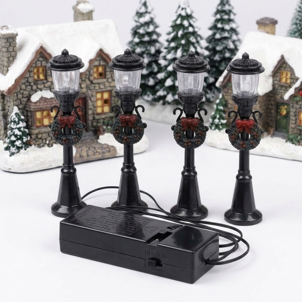 Four unlit black miniature street lights with wreaths and red bows stand before snowy model cottages, with connecting wires and a battery box.