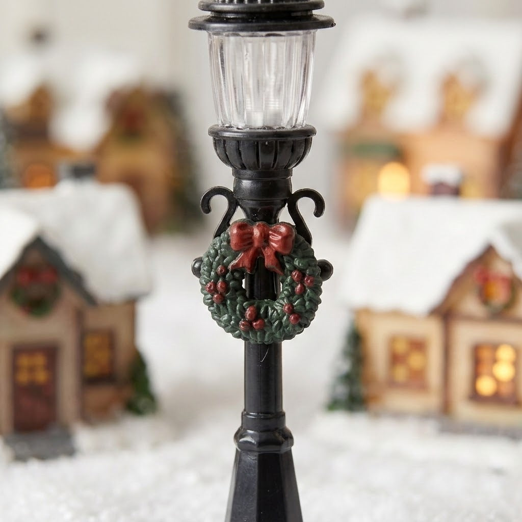 A black plastic miniature street light has a clear ribbed lantern, green wreath, and red bow against blurred snowy village houses.