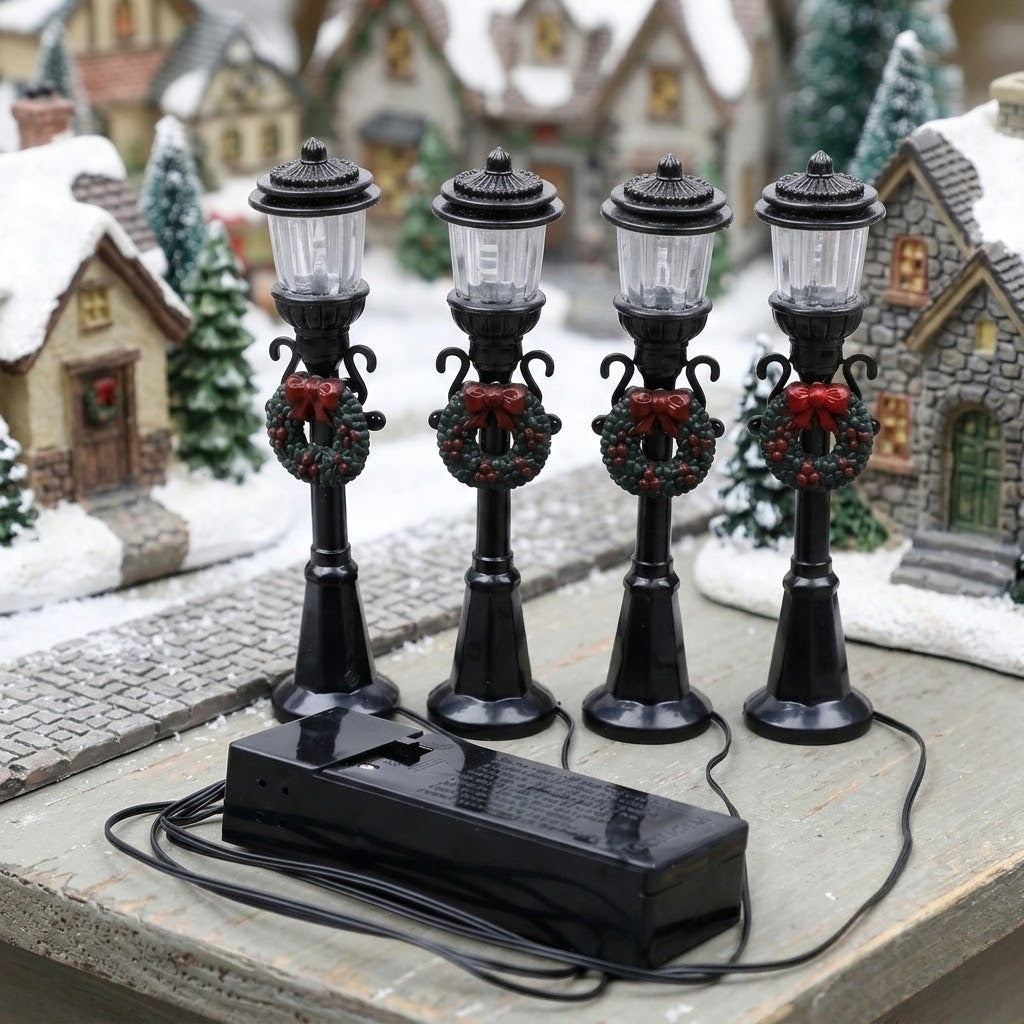Four unlit black miniature street lights with wreaths and red bows stand on wood beside snowy model houses and a wired battery box.