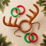 Ring toss game with brown inflatable antler headgear and four red and green rings on pale wood, framed by evergreen branches and string lights.