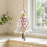A bell-shaped glass suncatcher with rainbow reflections and a pointed drop hangs beside a sunlit window, with a blurred plant behind.
