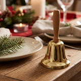 A gold hand bell with a wooden handle stands on a wooden dining table among plates, candles, and evergreen decorations.