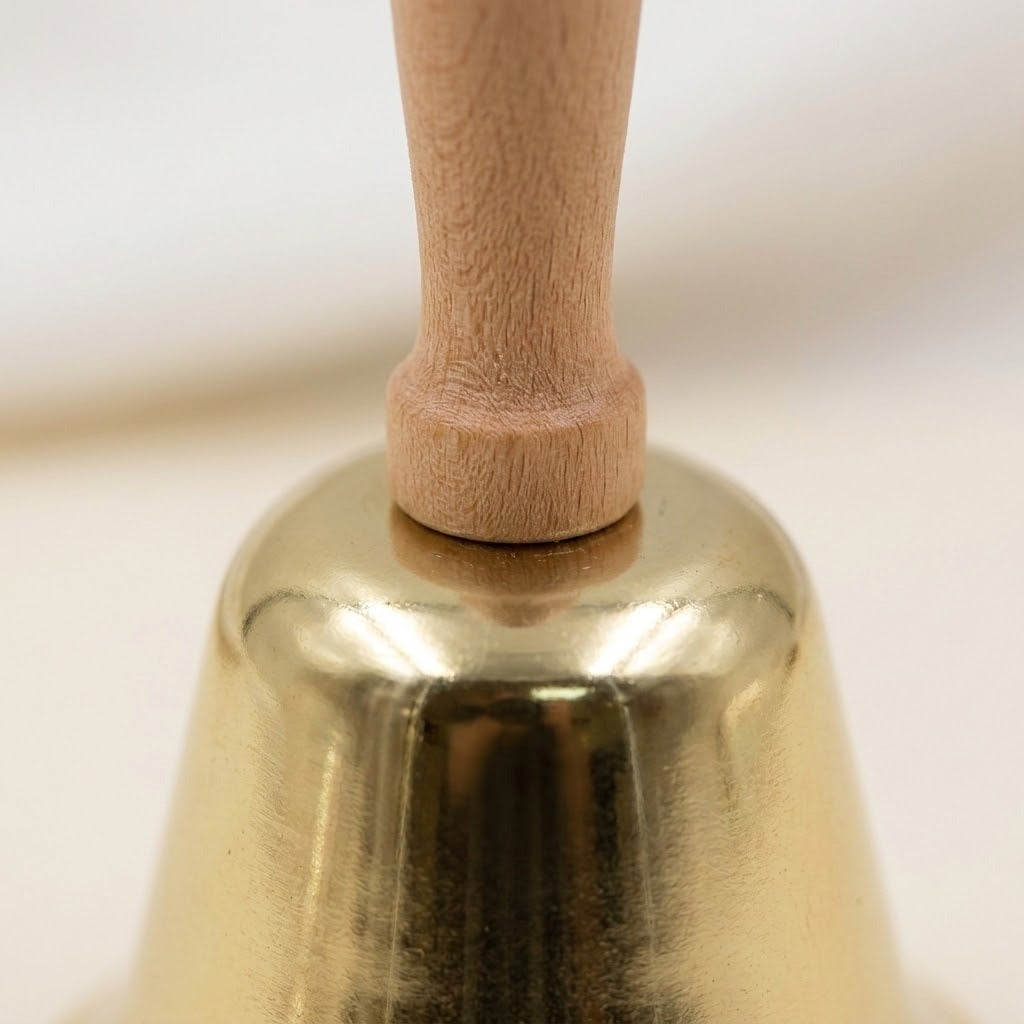 A close-up of the gold hand bell shows its wooden handle grain and reflective metal body against a cream background.