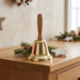 A gold hand bell with a wooden handle stands on a wooden cabinet beside evergreen branches, red berries, and a white star.