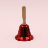 A shiny red iron hand bell with a light wooden handle stands upright against a pale pink background.