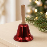 A glossy red hand bell with a wooden handle stands on a pale tabletop beside evergreen branches with blurred Christmas lights behind.