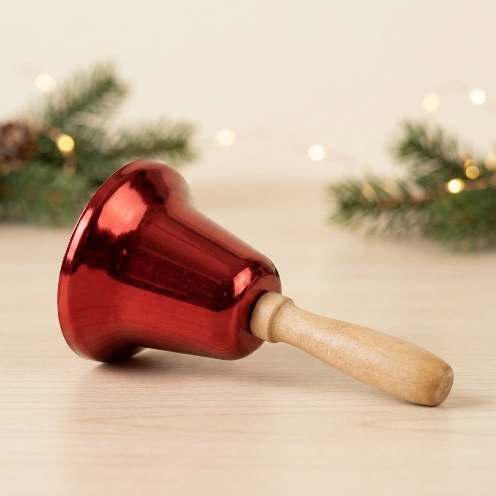 A glossy red hand bell lies sideways on pale wood, its wooden handle pointing right with evergreen sprigs behind it.