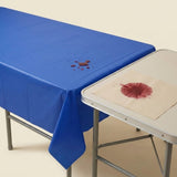 Reddish liquid beads on a blue tablecloth beside an uncovered folding table with a reddish spill on its surface.