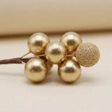 A gold foam berry pick has five smooth berries and one glittered berry on a brown stem against a beige background.