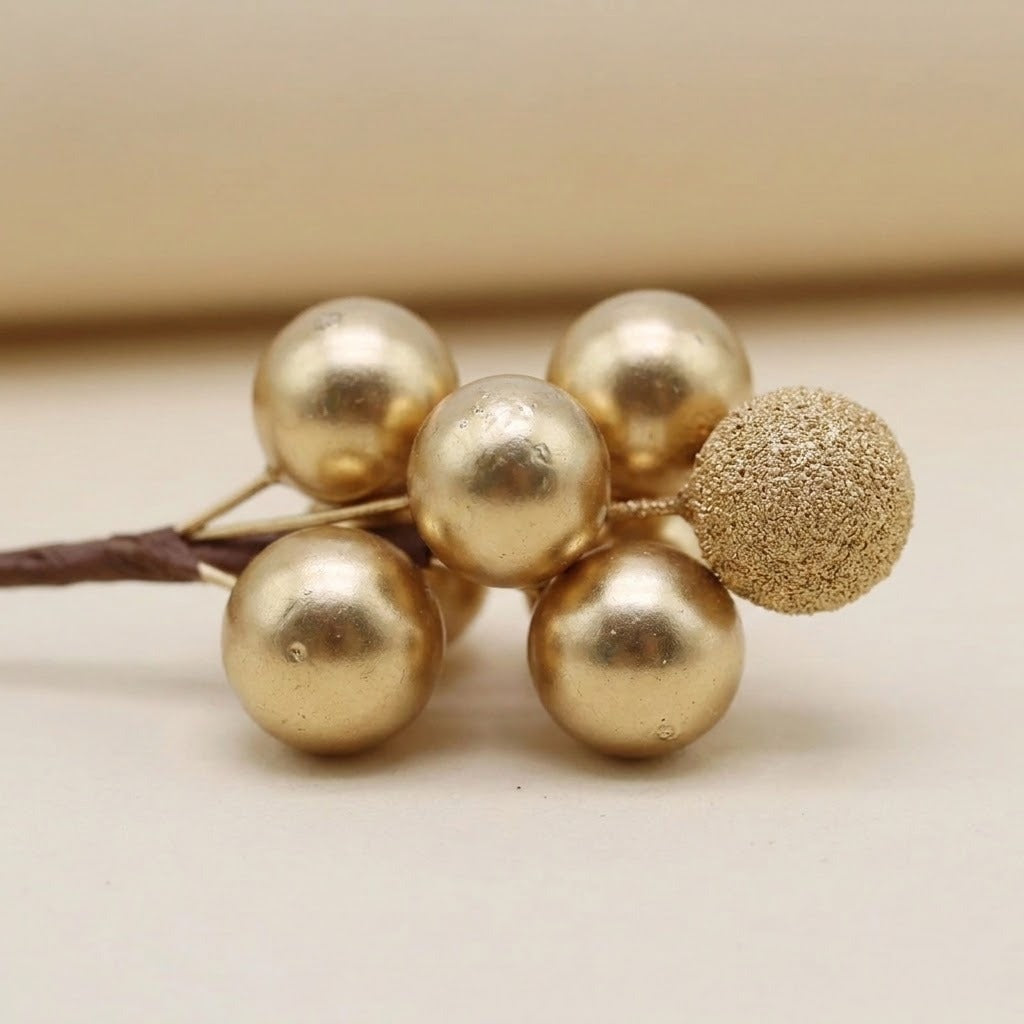 A gold foam berry pick has five smooth berries and one glittered berry on a brown stem against a beige background.